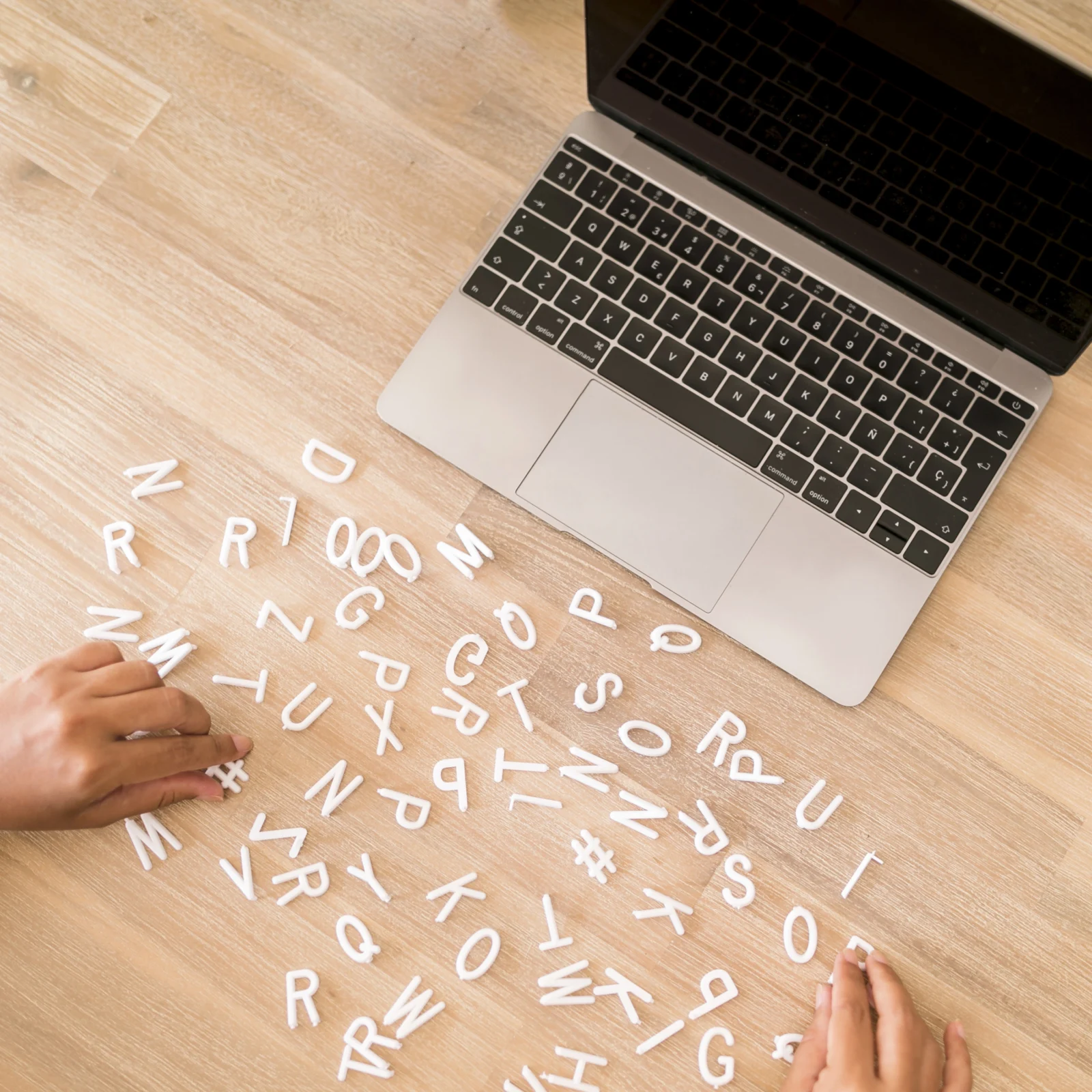 Ordinateur portable sur bureau avec lettres alphabétiques éparpillées manipulées à la main, illustrant la saisie de données, la rédaction et la gestion de contenu numérique