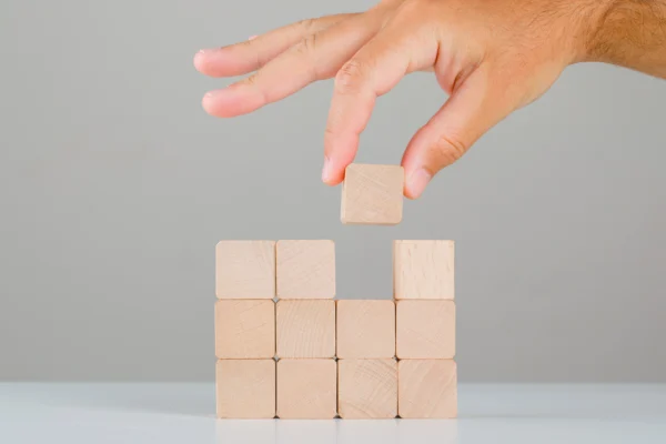 Main empilant des cubes en bois pour construire une structure, illustrant la mise en place d’une stratégie, l’organisation des processus et la construction d’un projet en entreprise