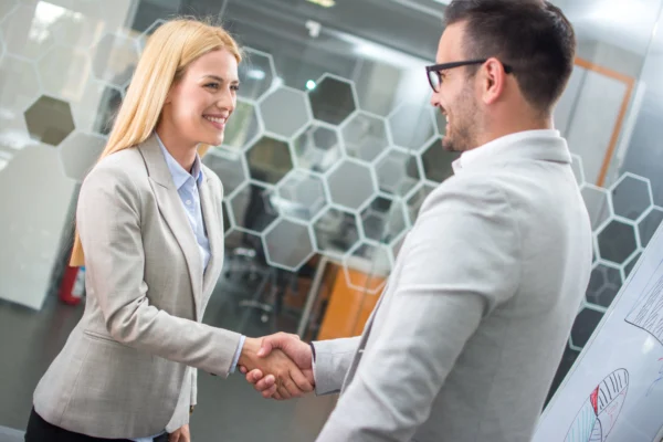 Deux professionnels en tenue formelle se serrant la main lors d’un échange en entreprise devant un tableau et un motif graphique, illustrant un accord commercial, un partenariat et la collaboration professionnelle
