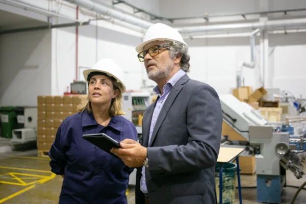 Deux professionnels équipés de casques de sécurité analysant une installation industrielle avec tablette en main, illustrant l’audit, le contrôle qualité et la gestion de production en usine