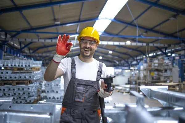Opérateur en usine équipé de casque et gants de sécurité faisant un geste OK tout en tenant une pièce mécanique, illustrant la qualité de production et le contrôle industriel