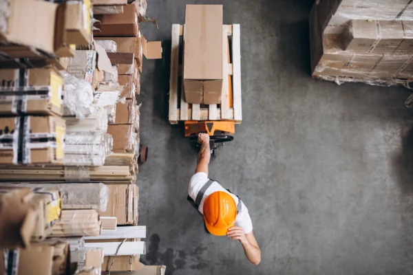 Vue de dessus d’un opérateur en entrepôt déplaçant une palette de cartons avec un transpalette, illustrant la logistique, la gestion des stocks et la préparation de commandes